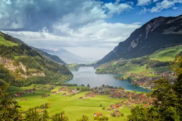 Panoramic view of a Swiss valley with a lake, rolling green hills, and mountain peaks.