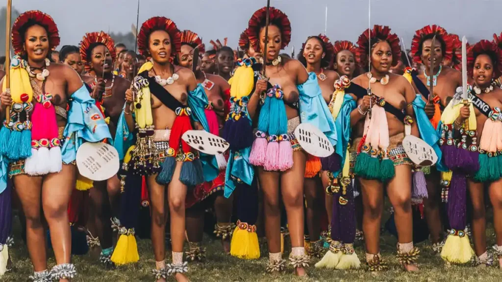 Group of women in vibrant traditional Eswatini attire, many holding spears, participating in a cultural celebration.